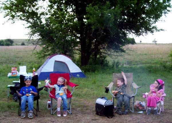 Four children sitting in camp chairs by a tent at dusk, eating together in a Kansas field