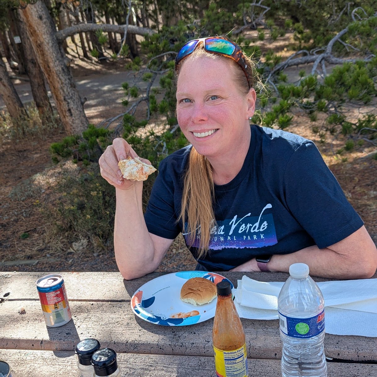 Marilee at a picnic table under pine trees, eating a crab sandwich on a road trip through Rocky Mountain National Park